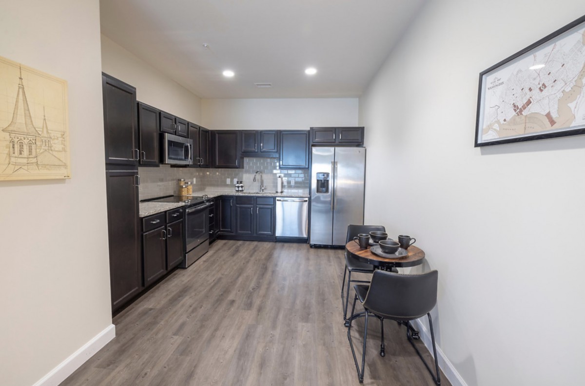 A modern kitchen with dark cabinetry and stainless steel appliances, featuring a small dining area with two chairs on the side.