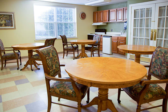An empty dining area with wooden tables and chairs, adjacent to a small kitchen.