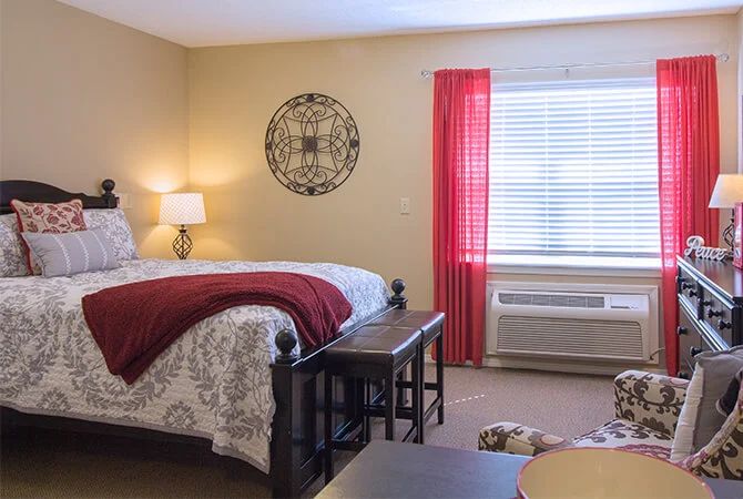A cozy bedroom with a neatly made bed, decorative wall art, and a window with red curtains.