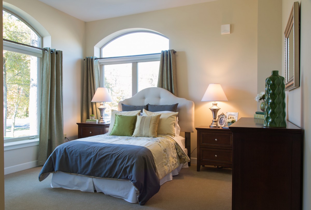 A cozy bedroom featuring a neatly made bed with multiple pillows, flanked by two bedside tables and lamps, with curtains framing a large window.