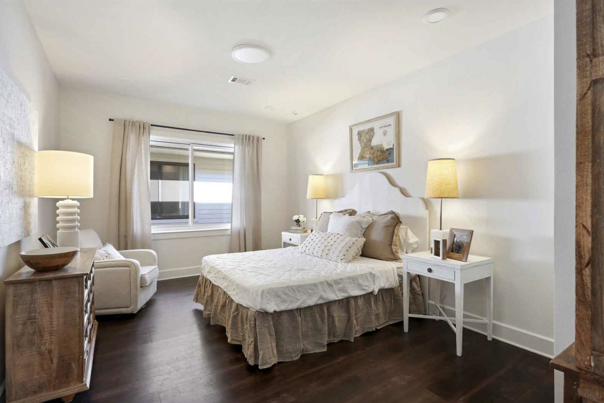 A cozy bedroom with modern white decor, featuring a bed, lamps, and a comfortable chair by the window.