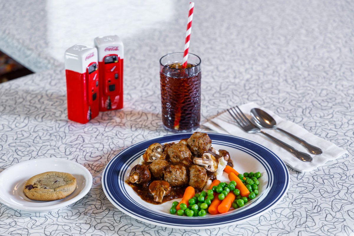 A plate of meatballs with peas and carrots, served with gravy and a soft drink in a retro diner setting.
