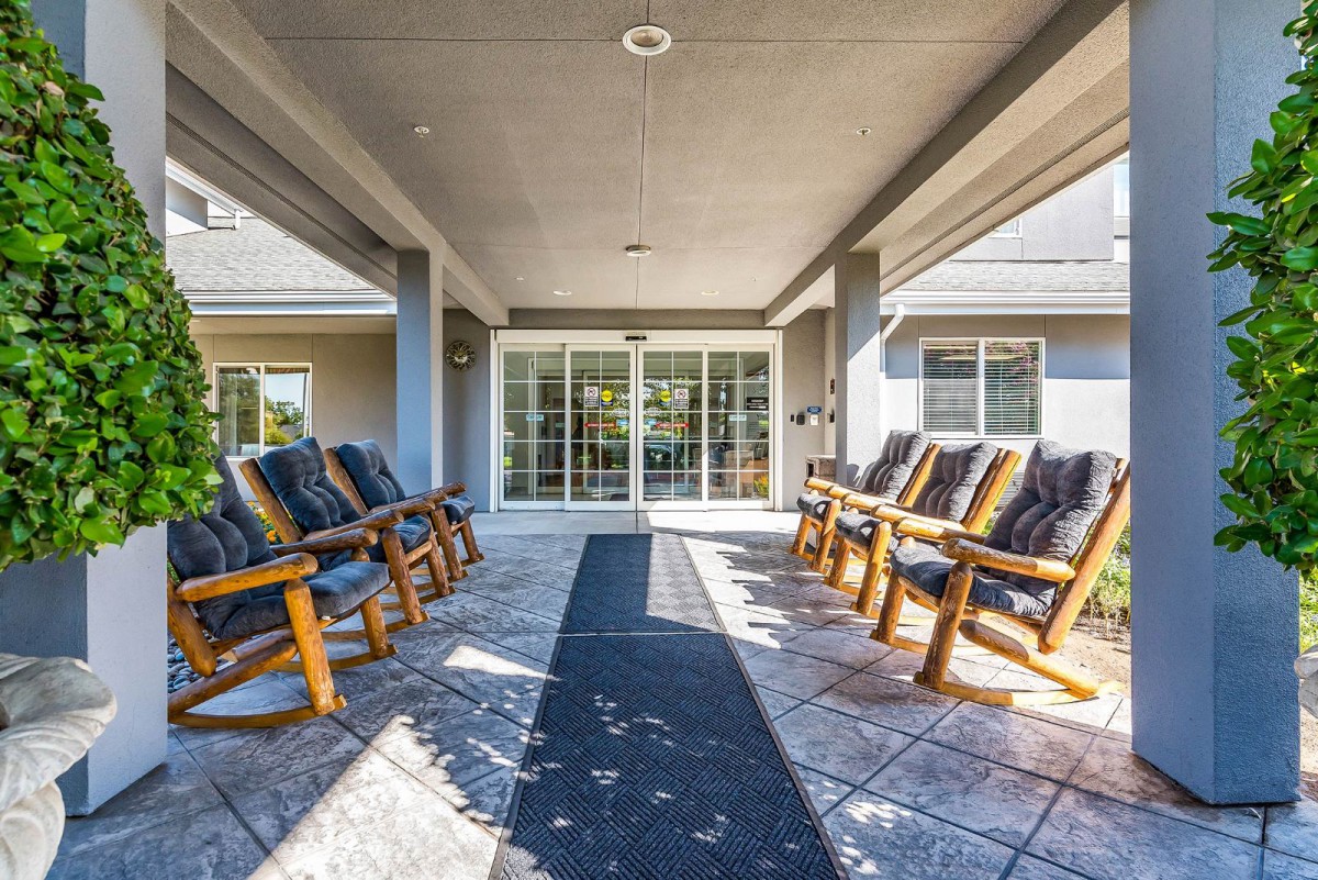 A sunlit patio with rows of wooden rocking chairs facing each other, under a covered roof and adjacent to glass doors.