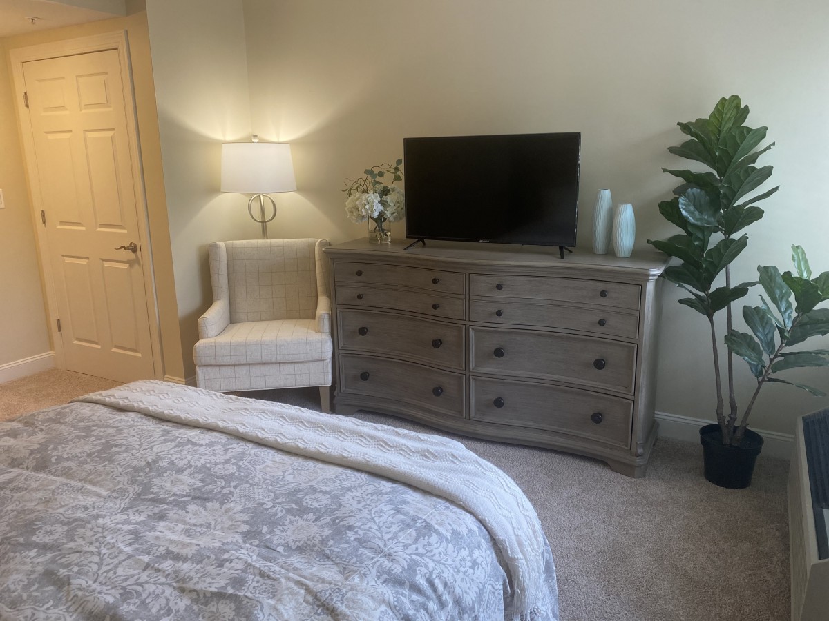 A cozy bedroom with a dresser, TV, lamp, and chair beside a bed.
