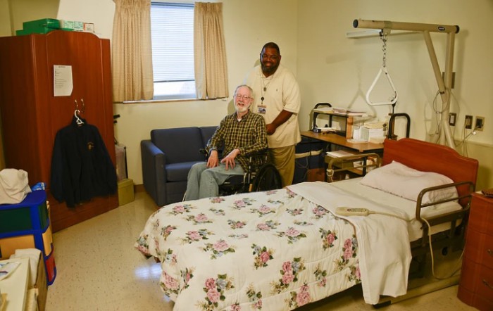 A caregiver stands beside a patient in a wheelchair in a hospital room.