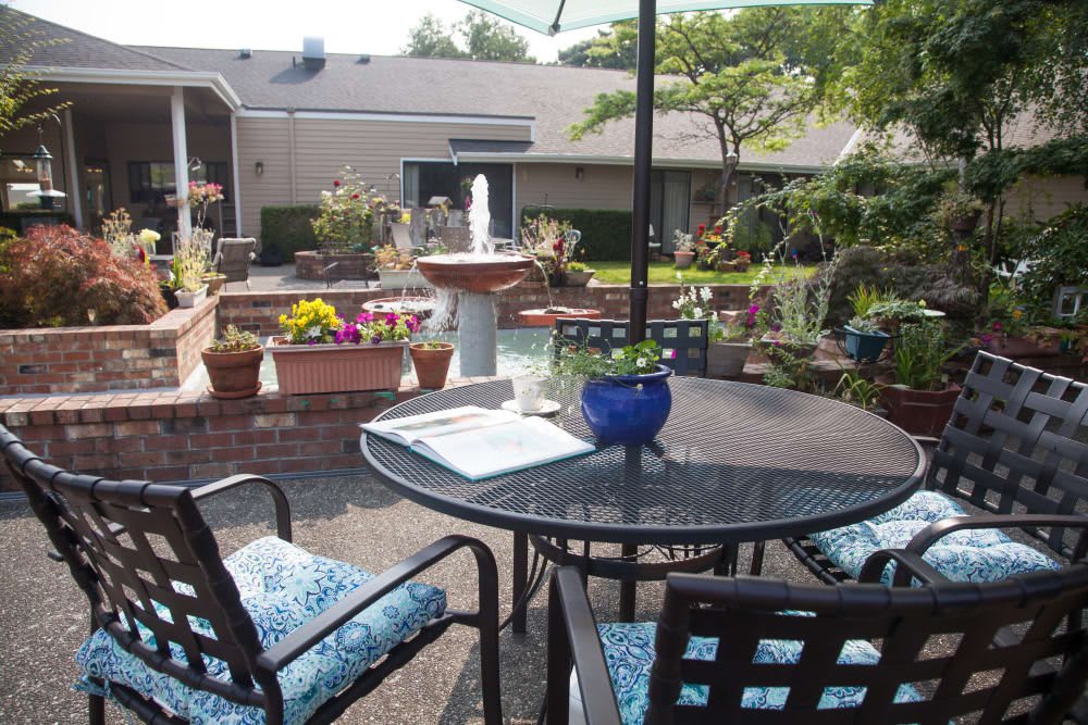 A cozy outdoor patio area with a glass table, cushioned chairs, and various flowers surrounding a fountain.