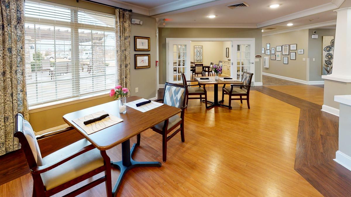A cozy dining area with neatly arranged tables and chairs, wooden flooring, and large windows letting in natural light.