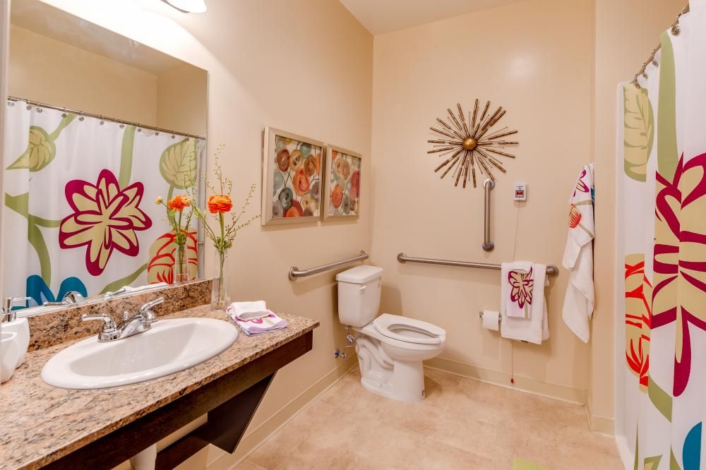 A well-decorated bathroom featuring floral artwork, a sink with flowers, and a toilet next to safety handrails.