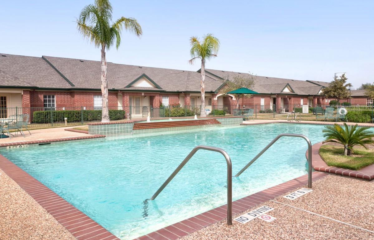 An outdoor swimming pool in a residential area, surrounded by palm trees and brick buildings.