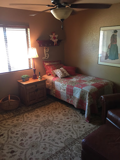 A cozy bedroom featuring a single bed dressed in a colorful quilt, accompanied by a nightstand and lamp, with natural light flowing through a window.