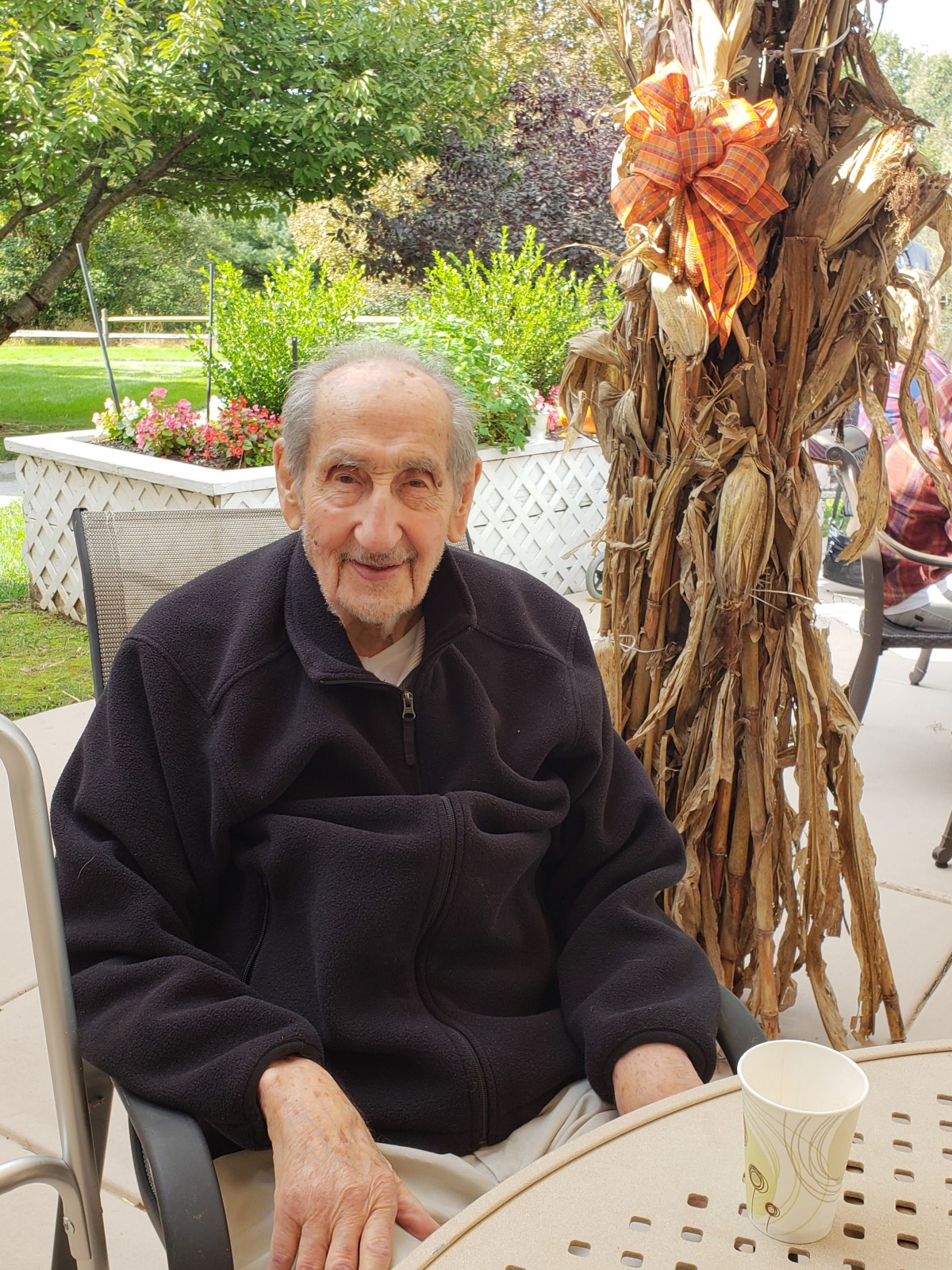 An elderly man sits outdoors, smiling with autumn decorations nearby.
