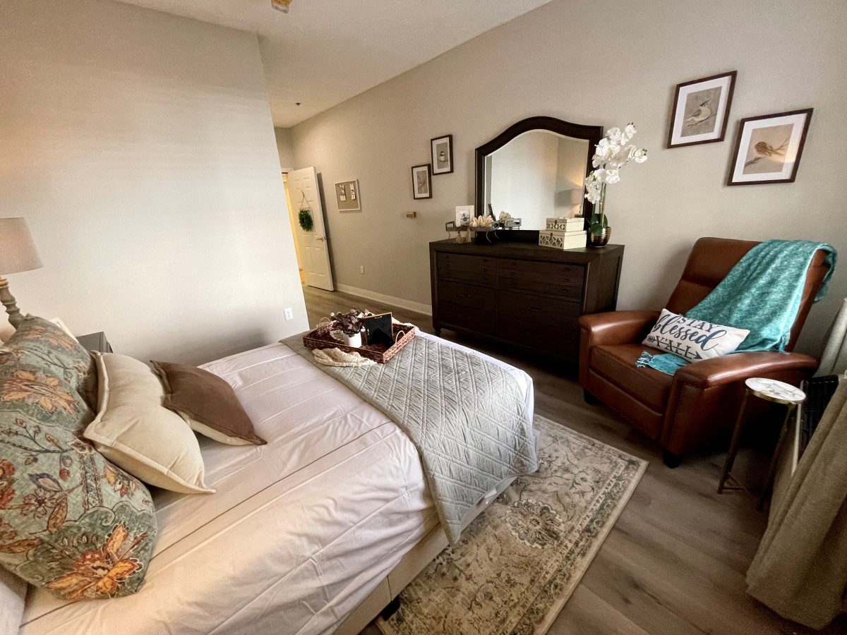 A cozy bedroom with a neatly made bed, an armchair, and a dresser with a mirror.