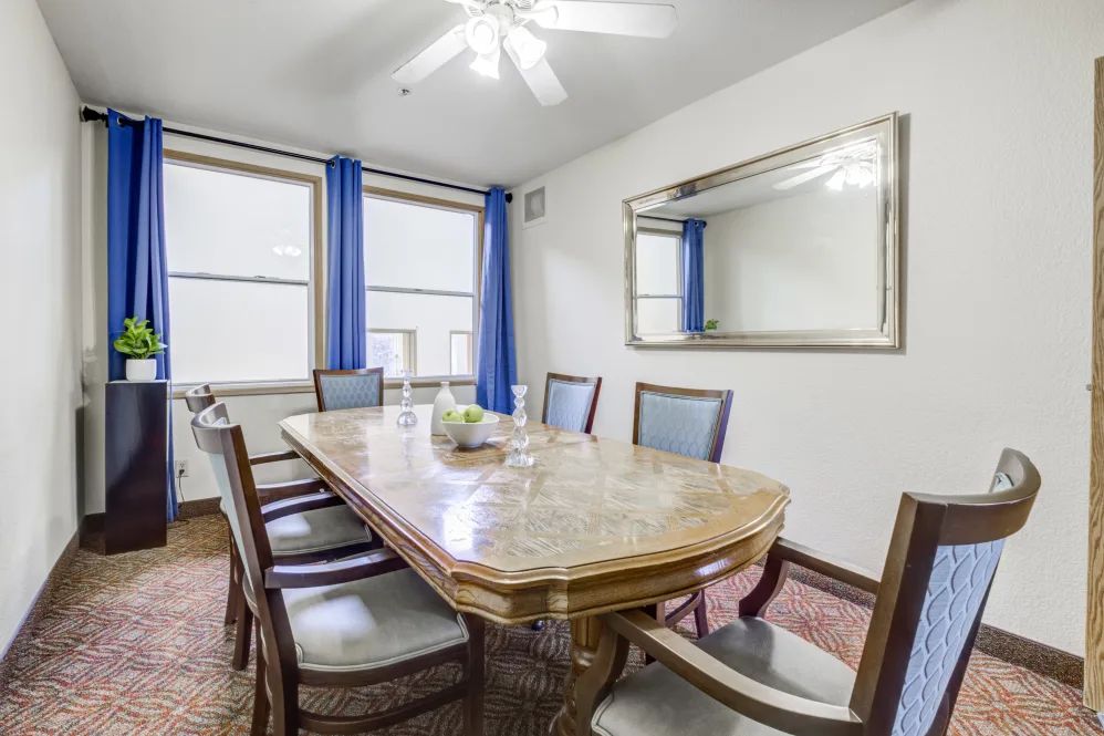 A cozy dining room with a wooden table, surrounded by chairs, featuring blue curtains and a large mirror.