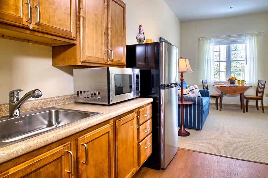 A compact kitchen area with wooden cabinets featuring a microwave, refrigerator, and sink, adjacent to a cozy living room.