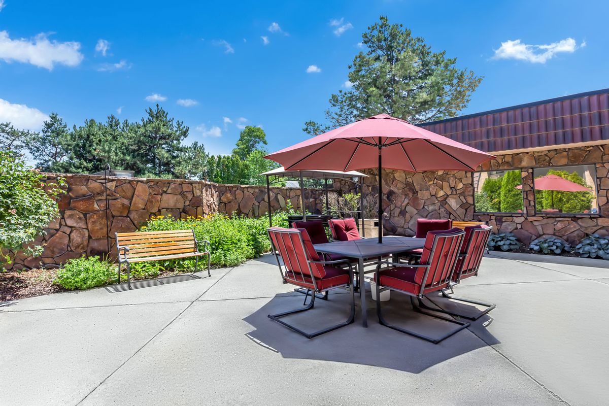 A sunny patio with a table and chairs set under a large red umbrella, surrounded by a garden.