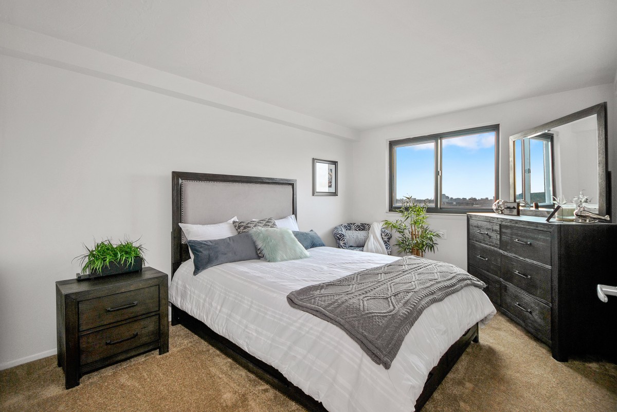 A minimalist bedroom featuring a neatly made bed, a dresser, and potted plants with natural light coming through the window.