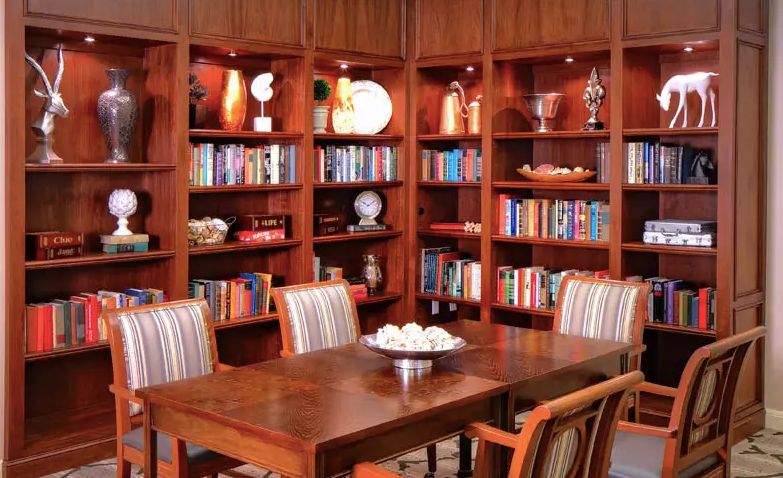 A cozy library with wooden bookshelves and a dining table surrounded by chairs, adorned with books and decorative items.