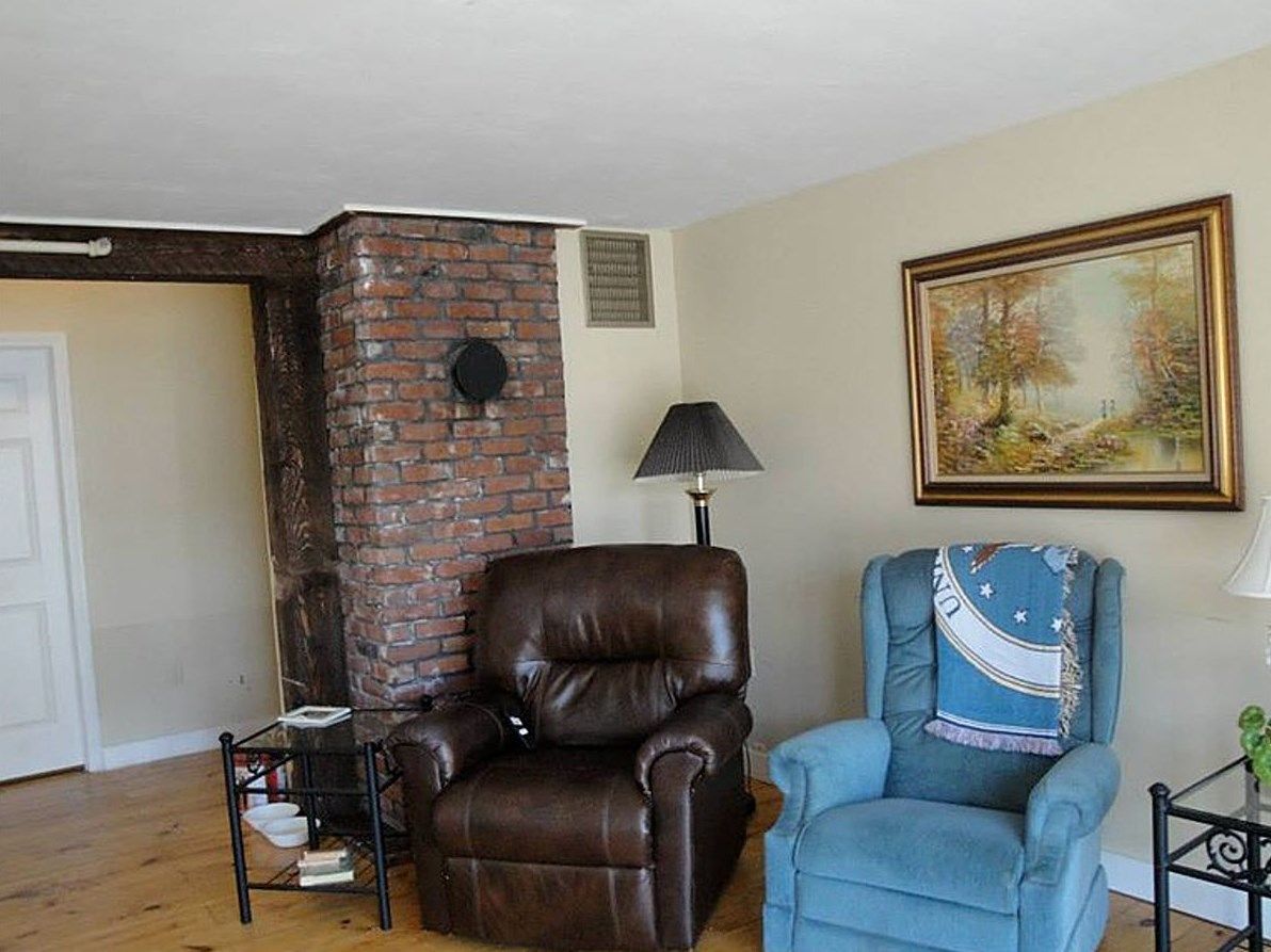 A cozy living room with a brick wall, featuring a brown leather armchair and a blue armchair next to a lamp and painting.