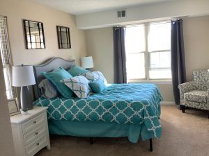 A cozy bedroom with a teal and white bedspread, multiple pillows, and two windows with dark curtains.