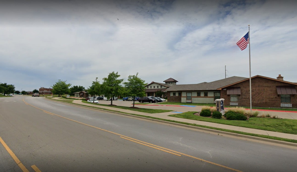 A street view showing a large building with a parking lot, surrounded by trees and a sidewalk under a cloudy sky.