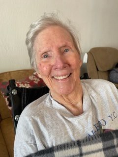 An elderly person smiling indoors, wearing casual clothing.