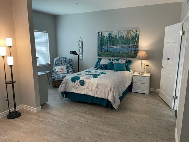 A cozy bedroom featuring a neatly made bed with teal accents, a decorative painting above the headboard, and a comfortable reading chair by the window.