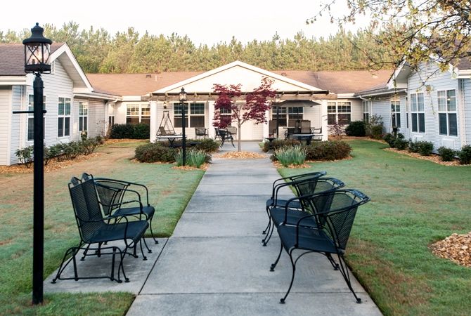 A peaceful outdoor area with chairs lining a pathway leading to residential buildings surrounded by lush landscaping.