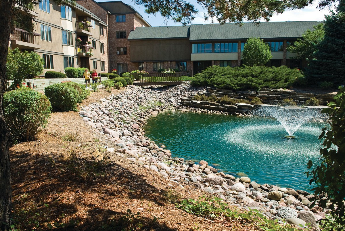 A tranquil pond with a fountain surrounded by lush landscaping near an apartment complex.