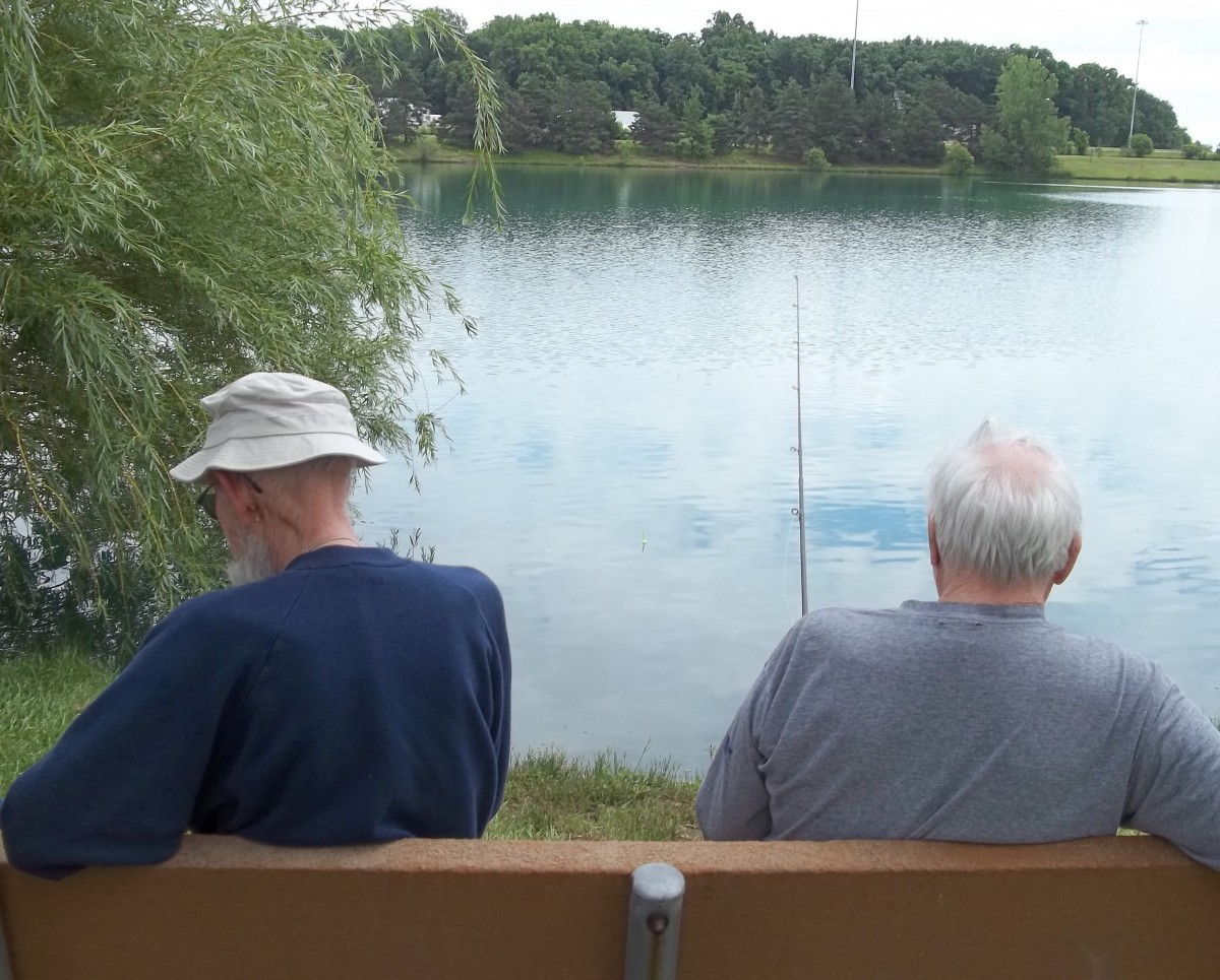 Two elderly men sitting on a bench, fishing by a tranquil lake.