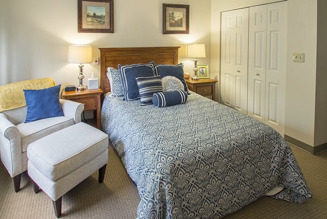 A cozy bedroom with a neatly made bed, featuring a blue patterned quilt and several pillows. Two bedside lamps illuminate the room, which includes framed artwork on the walls.