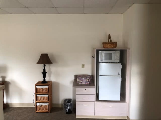 A room with a lamp on a small drawer unit and a cabinet housing a microwave and refrigerator.