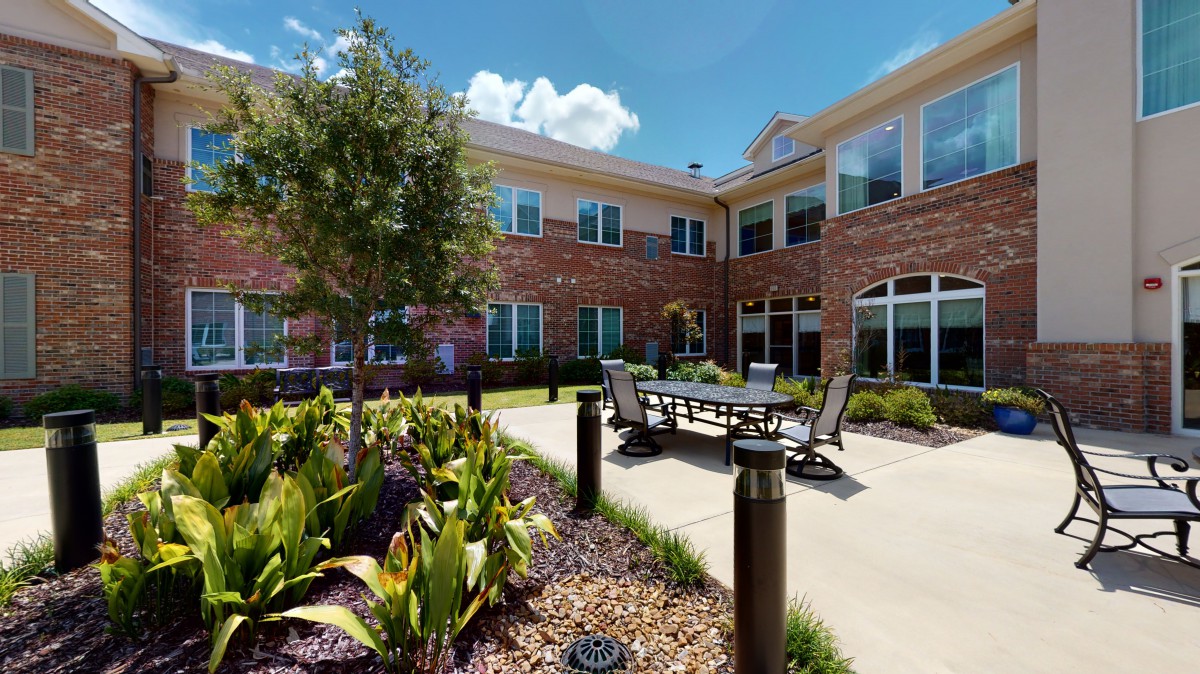 A sunny courtyard with outdoor seating and a landscaped garden in front of a brick building.