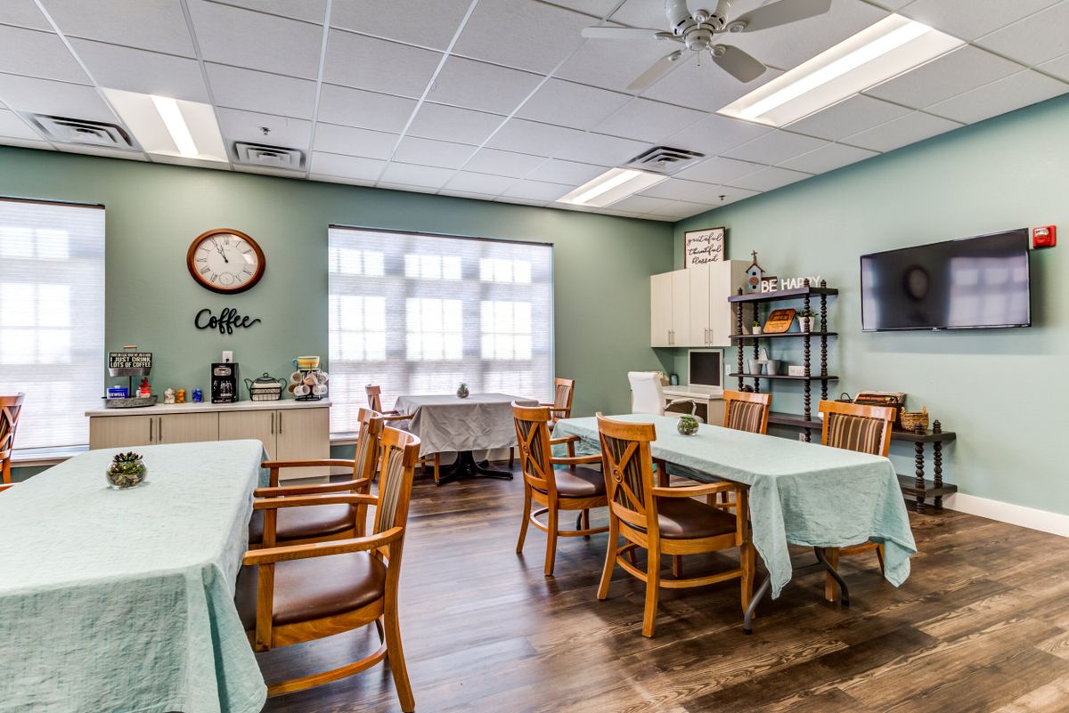 A cozy dining area with tables and chairs, decorated with a clock and natural light from large windows.