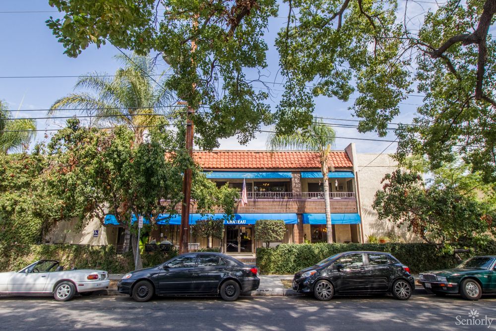 A street view featuring parked cars in front of a building surrounded by trees.
