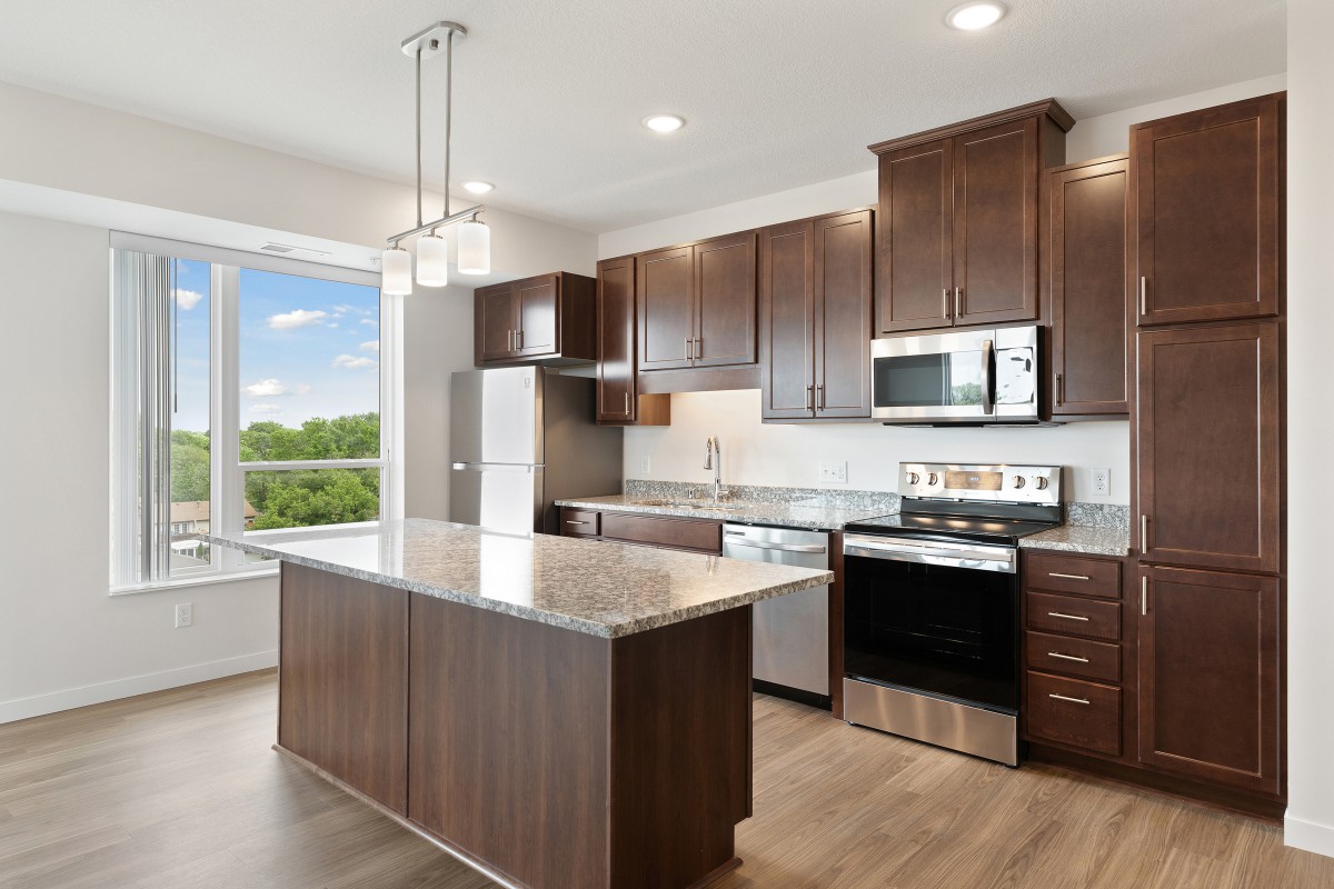 A modern kitchen with wooden cabinets and stainless steel appliances, featuring a central island and large window.