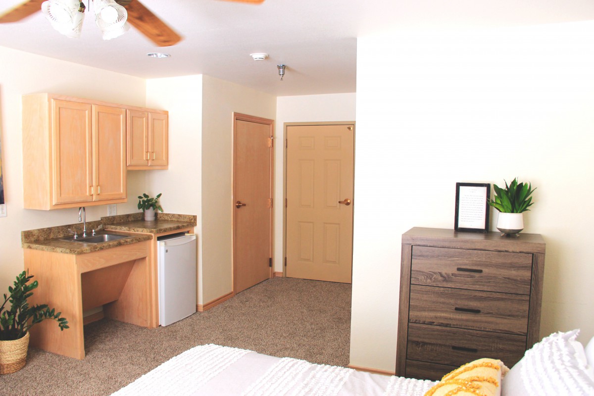 A cozy bedroom featuring a small kitchenette with a mini fridge, a ceiling fan, and a dresser.