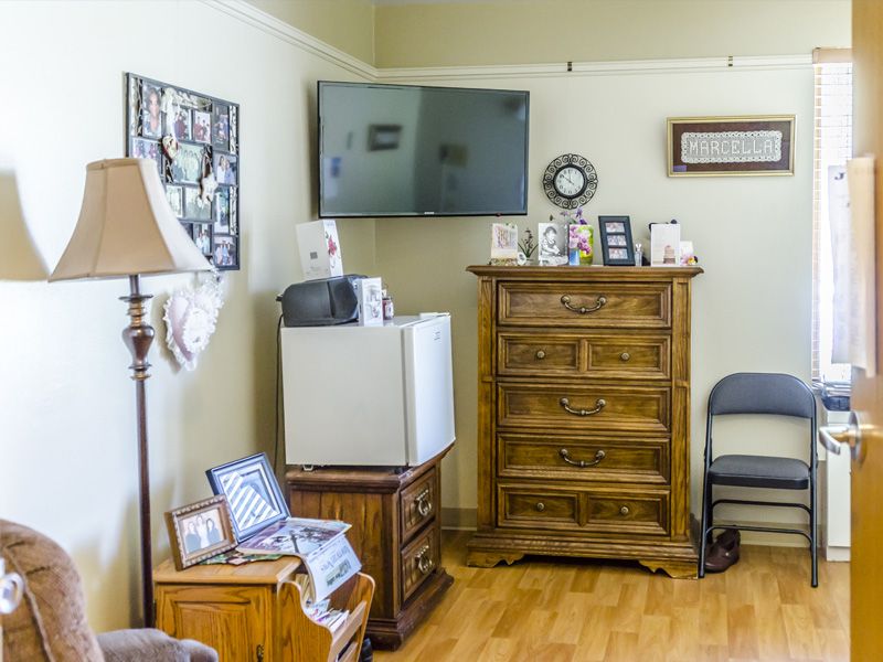 A cozy living room corner featuring a wall-mounted television, a wooden dresser, and various decorations including framed pictures and a lamp.