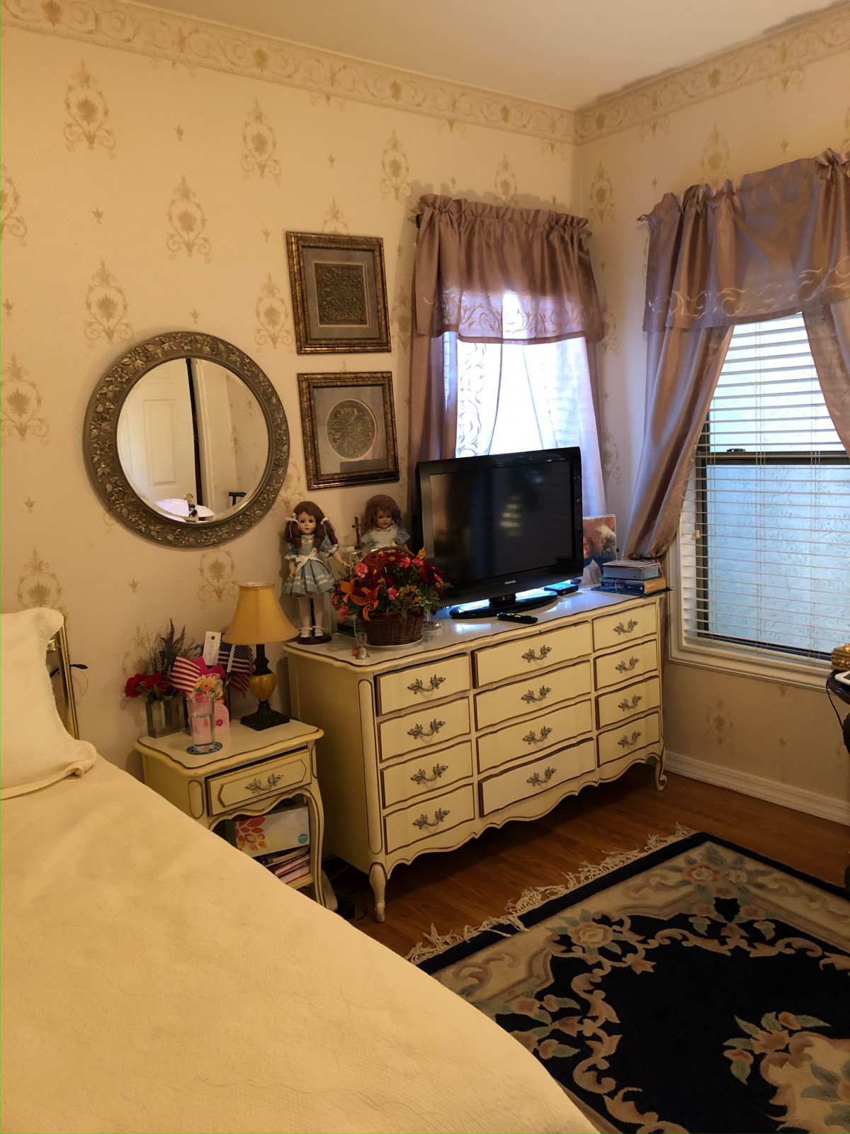 A cozy vintage-style bedroom featuring a dresser with a TV, decorative mirror, and elegant curtains.