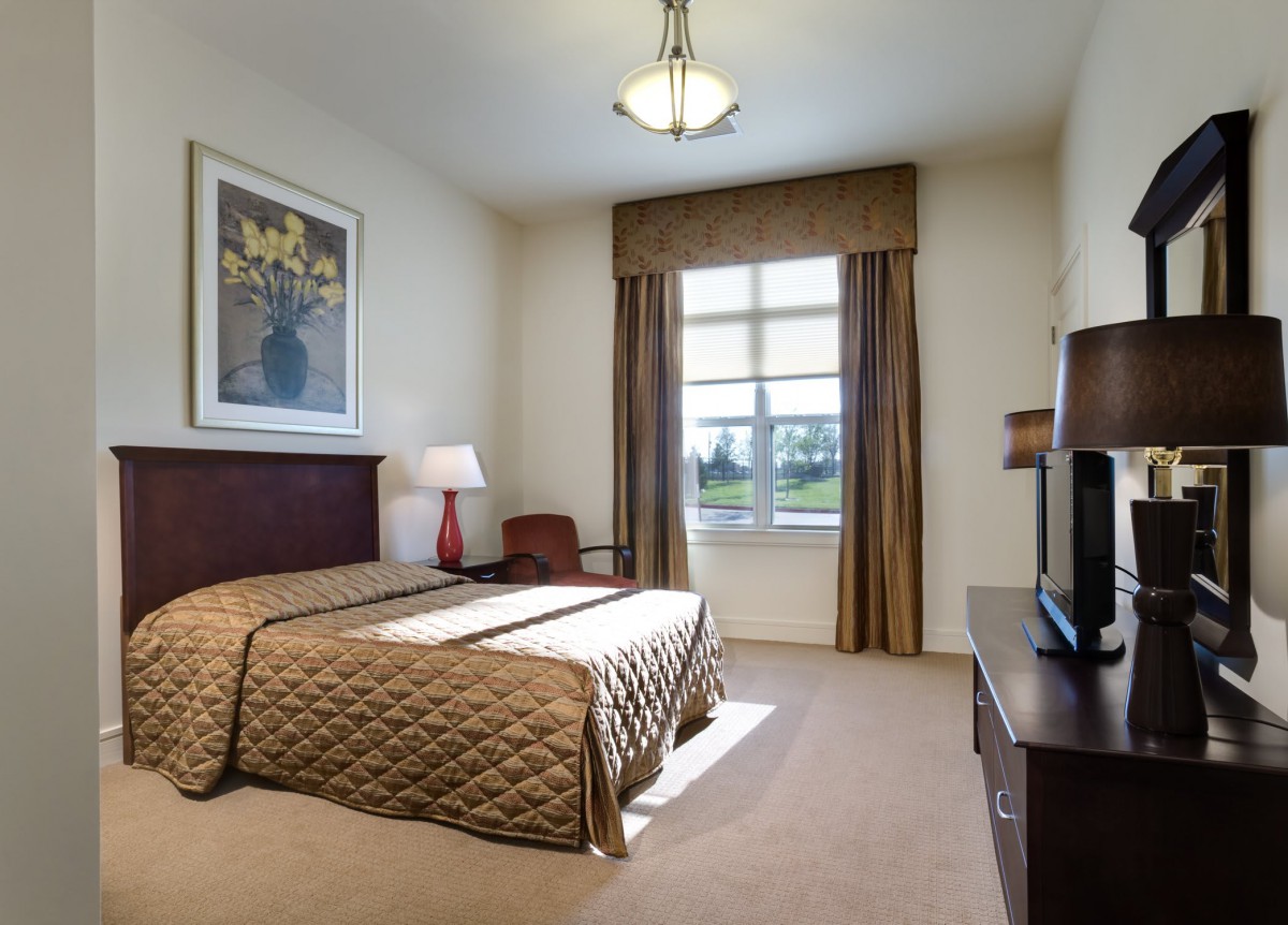 A well-lit hotel bedroom featuring a large bed, bedside lamp, and a window with curtains.