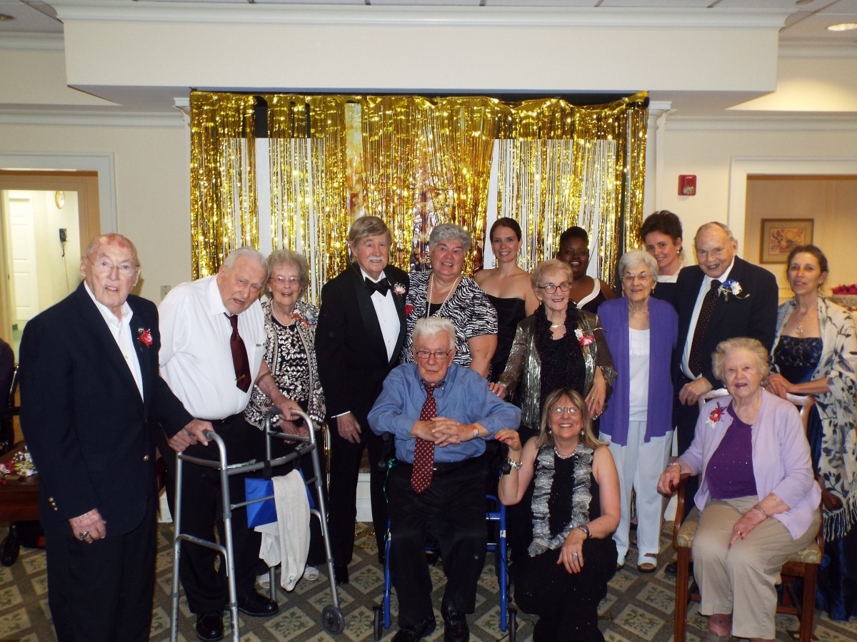 A group of people, mostly seniors, are gathered together dressed in formal attire for a celebration in front of a gold backdrop.