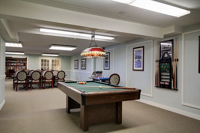 A well-lit basement game room features a billiard table, decorative lighting, and framed wall art.