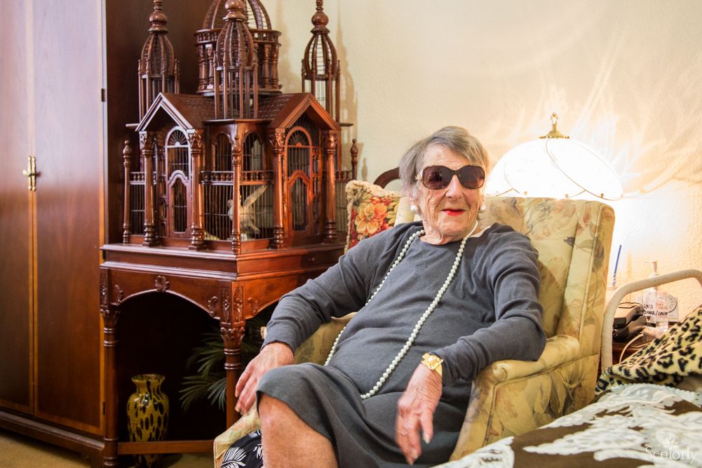 An elderly woman wearing sunglasses is seated in a living room next to an intricate wooden sculpture.