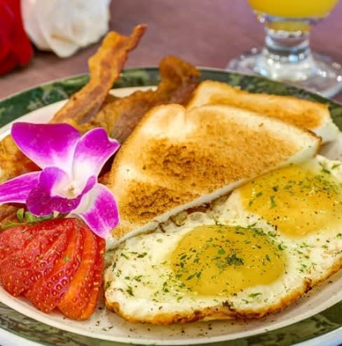 A delicious breakfast with eggs, toast, bacon, strawberries, and a flower garnish alongside a glass of orange juice.