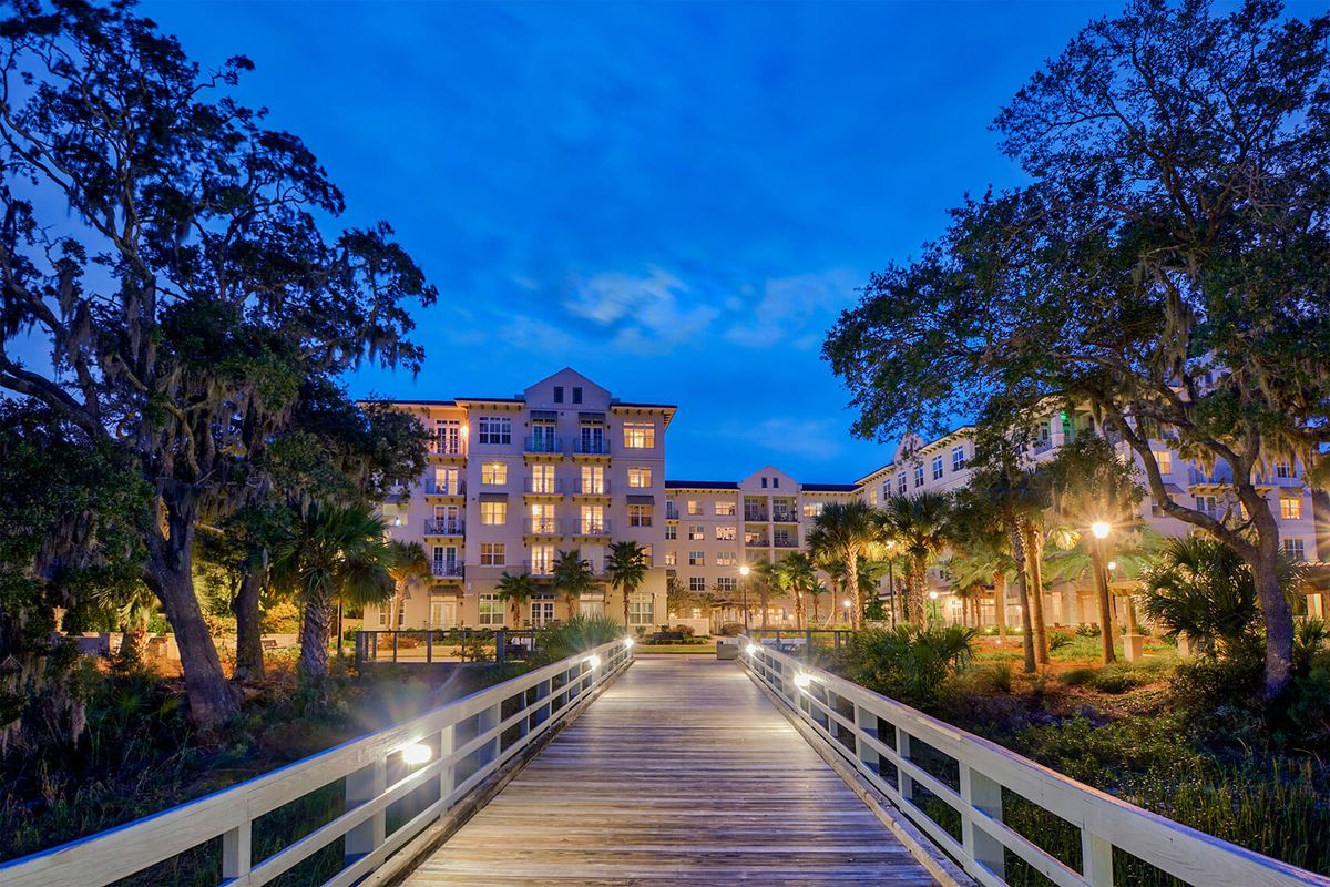 A luxurious hotel resort illuminated at night, with a well-lit bridge leading to the entrance surrounded by palm trees.