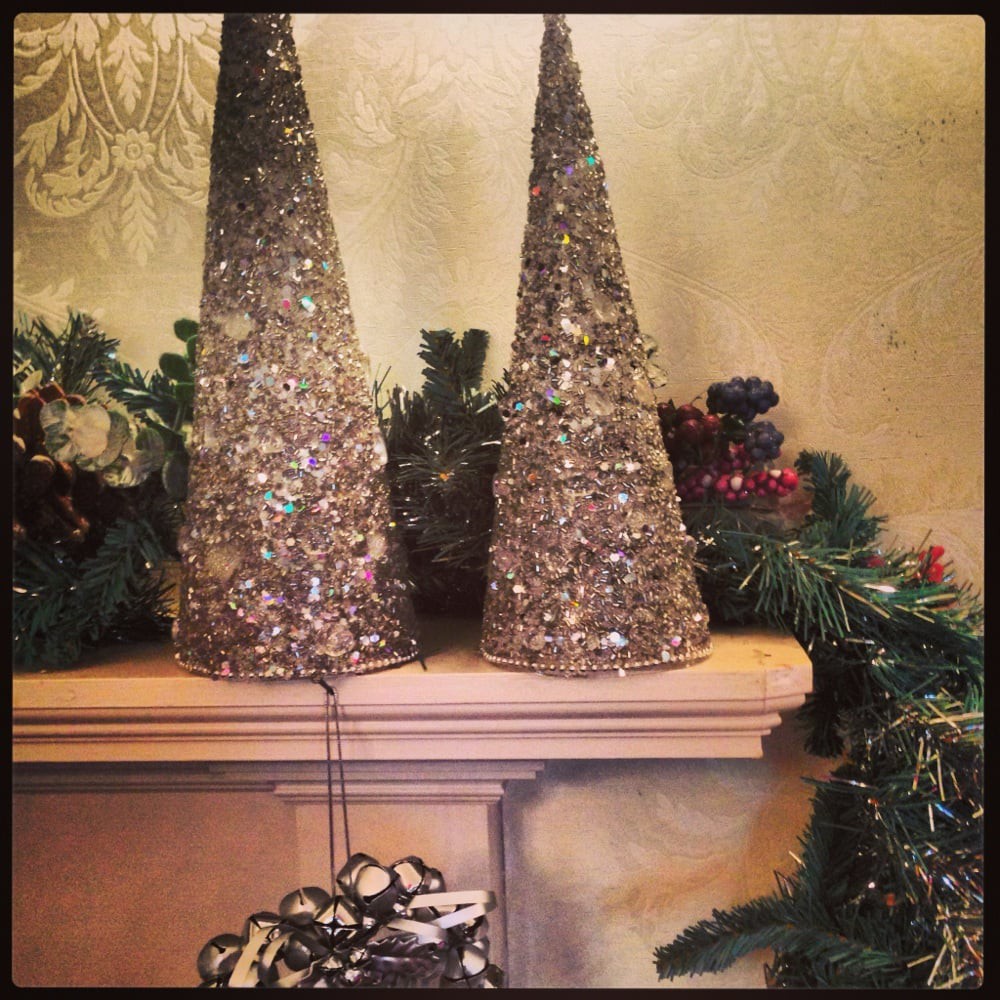 Festive holiday decorations with cone-shaped trees and garland on a fireplace mantel.
