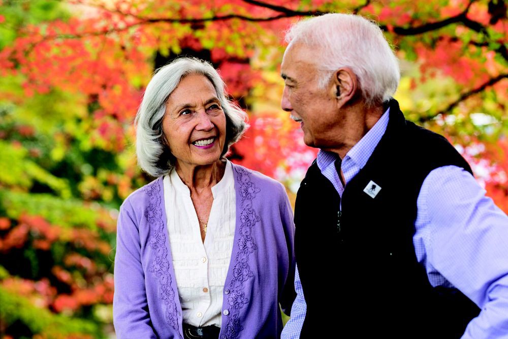 An elderly couple smiling at each other against a backdrop of autumn leaves.