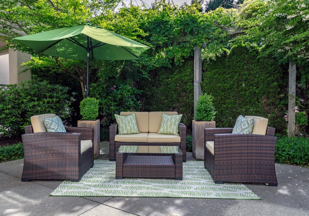 A cozy outdoor patio setup with wicker furniture, cushions, a table, and an umbrella surrounded by lush greenery.
