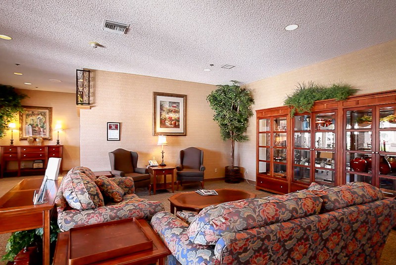 A cozy living room with floral sofas, chairs, and a coffee table, accented by indoor plants and warm lighting.