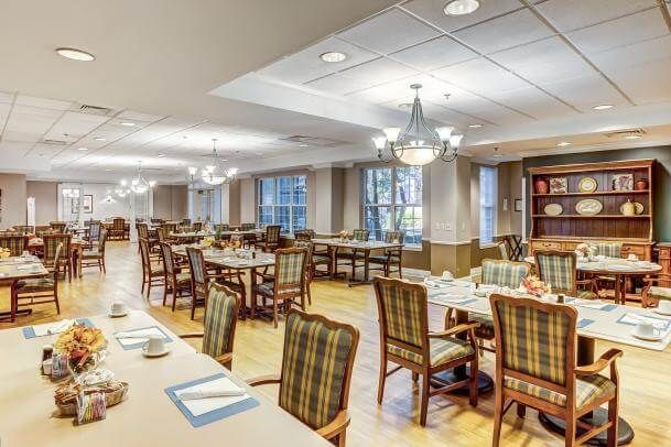 A spacious dining area with neatly arranged tables and chairs, illuminated by chandeliers and natural light from large windows.