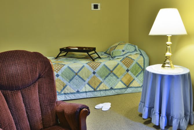 A cozy bedroom with a colorful quilt on the bed, a tray placed on top, and an armchair beside a lamp and side table.